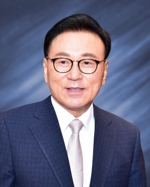 정장 차림의 남성이 카메라를 바라보며 미소 짓고 있는 모습이다. 배경은 흐릿한 색상으로 처리되어 있다.