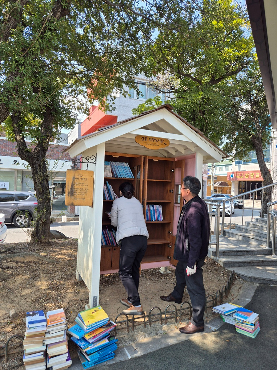 두 사람이 감천 작은 도서관에서 책을 고르고 있는 모습이다. 한 사람은 서가에서 책을 꺼내고 있으며, 다른 사람은 서가 옆에서 책을 살펴보고 있다.