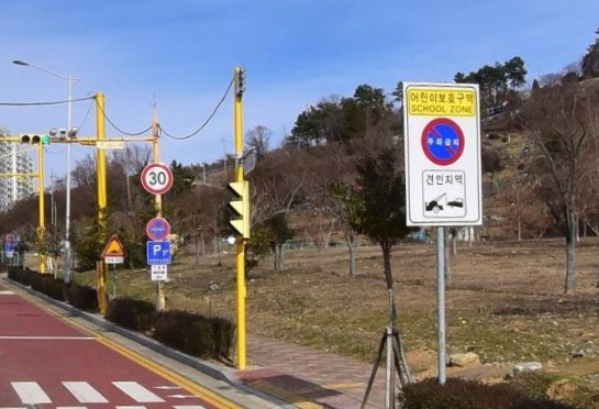 어린이 보호구역을 알리는 표지판이 있는 도로의 모습으로, 주변은 나무와 잔디가 있는 공터이다.