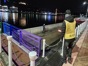 한 남성이 야경이 보이는 강가에서 안전을 위해 안내를 하고 있다. 배경에는 조명이 비치는 다리와 건물이 있다.