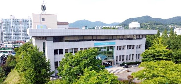 건물 외관이 보이는 장면으로, 주변에 나무와 산이 있는 모습이다. 건물 앞에는 계단과 출입구가 있다.