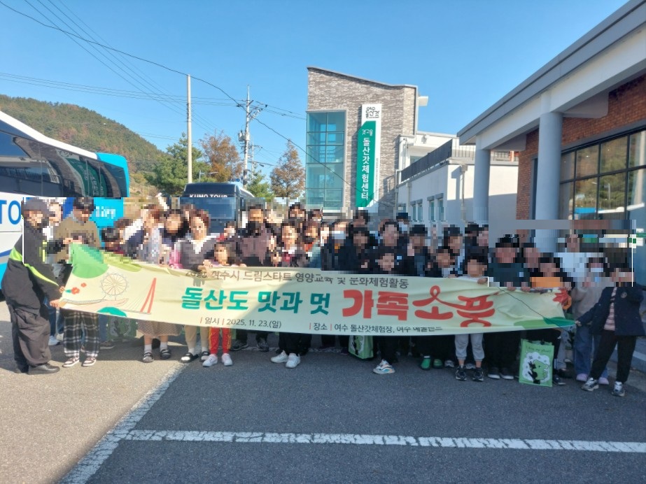 여수 드림스타트 영양교육 및 문화체험 활동 ‘돌산도 맛과 멋: 가족 소풍’ 행사에 참여한 아동과 가족 67명이 여수 돌산갓 체험장 앞에서 단체 사진을 찍고 있다. 참여자들이 ‘돌산도 맛과 멋 가족 소풍’ 현수막을 들고 있으며, 뒤편에는 돌산갓 체험센터 건물이 보인다