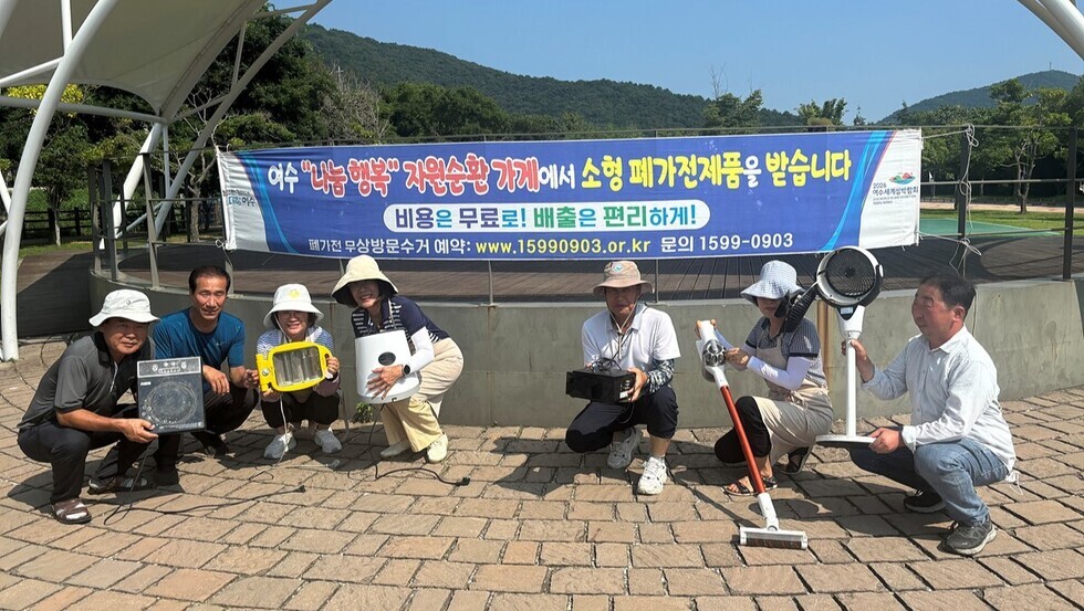 여수 <나눔 행복> 자원순환가게에서 소형 폐가전제품을 받습니다. 비용은 무료로! 배출은 편리하게! 폐가전 무상방문수거 예약 : www.15990903.or.kr 문의 1599-0903 [2026 여수세계섬박람회] 현수막이 걸려있고, 각 청소기, 난로, 선풍기 등 소형 폐가전제품을 들고 앉아 있는 7명의 사람들을 찍은 모습