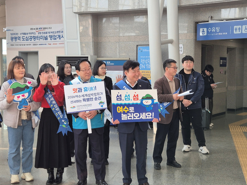 여수세계섬박람회를 홍보하는 행사에서 여러 사람이 모여 손을 흔들며 포즈를 취하고 있다. 이들은 여수로 관광을 유도하는 현수막을 들고 있다.