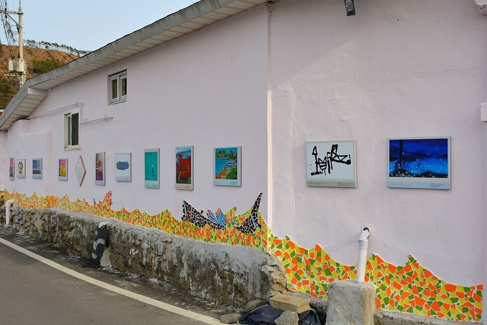 분홍색 벽에 여러 개의 그림이 걸려 있는 장면으로, 벽 아래에는 다채로운 색상의 모자이크가 장식되어 있다.