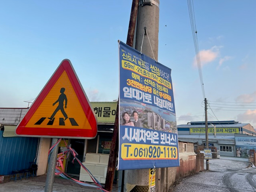 신도시 선택을 홍보하는 현수막과 보행자 신호등이 있는 거리 풍경. 현수막에는 주택형 정보와 연락처가 적혀 있다.