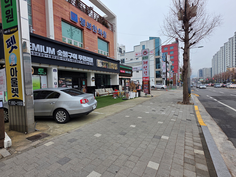 명보한의원과 PREMIUM 숯불구이 무한리필 식당이 있는 거리 풍경으로, 식당 앞에는 소고기와 오징어갈비를 제공하는 메뉴가 보인다.