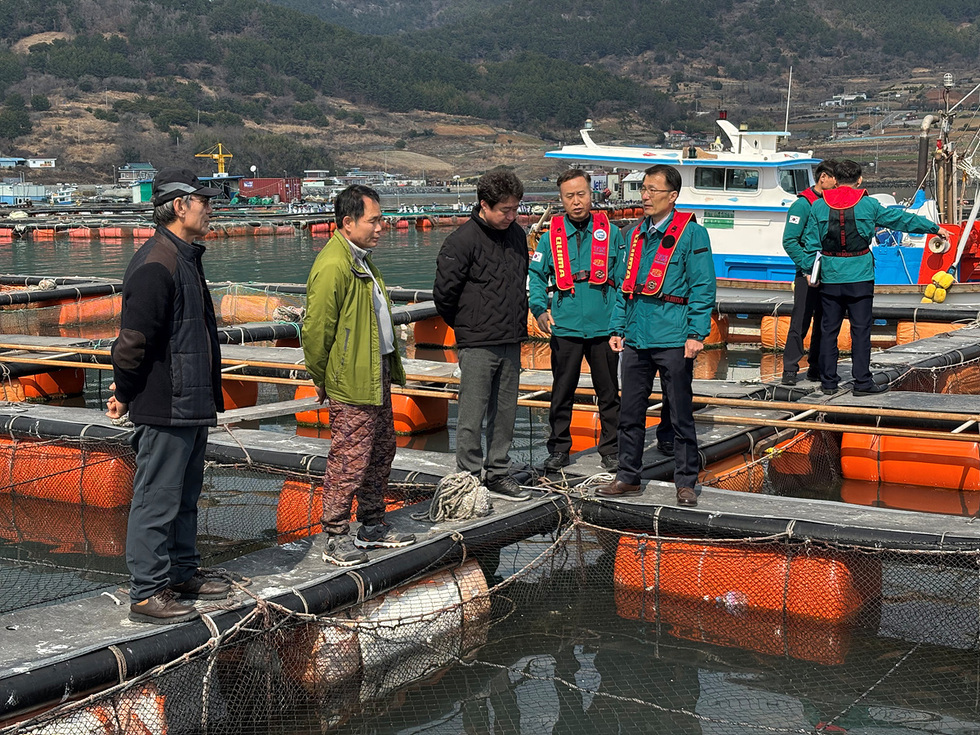 어업 현장에서 여러 사람이 수산 양식장에 대해 논의하고 있는 장면이다. 이들은 안전 장비를 착용하고 있으며, 배경에는 어선과 양식장이 보인다.