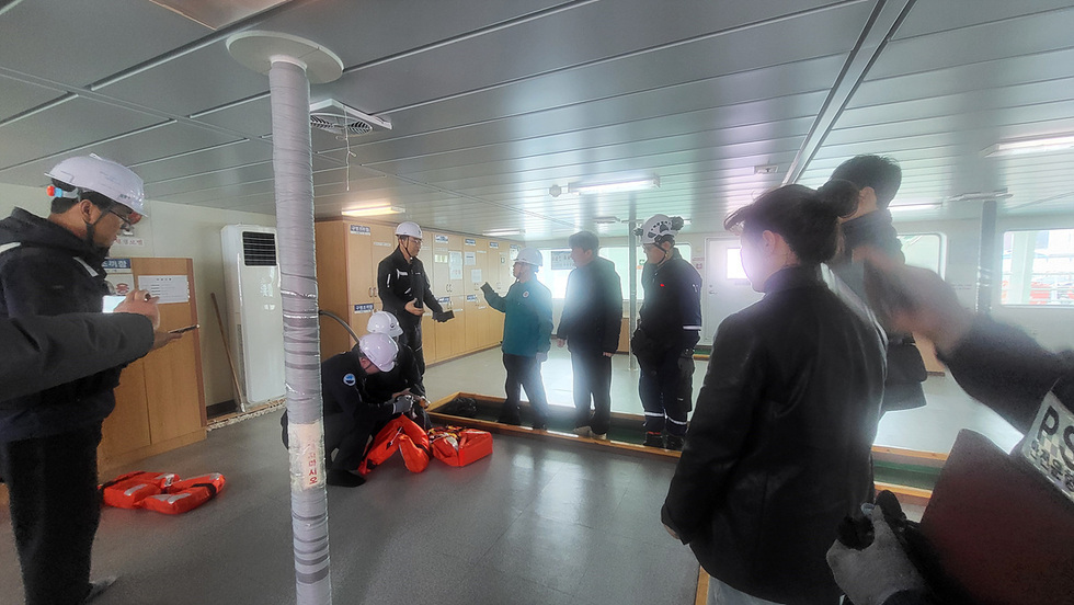 안전교육을 받는 여러 명의 사람들이 실내에서 훈련을 진행하고 있으며, 한 사람이 장비를 설명하고 있습니다.