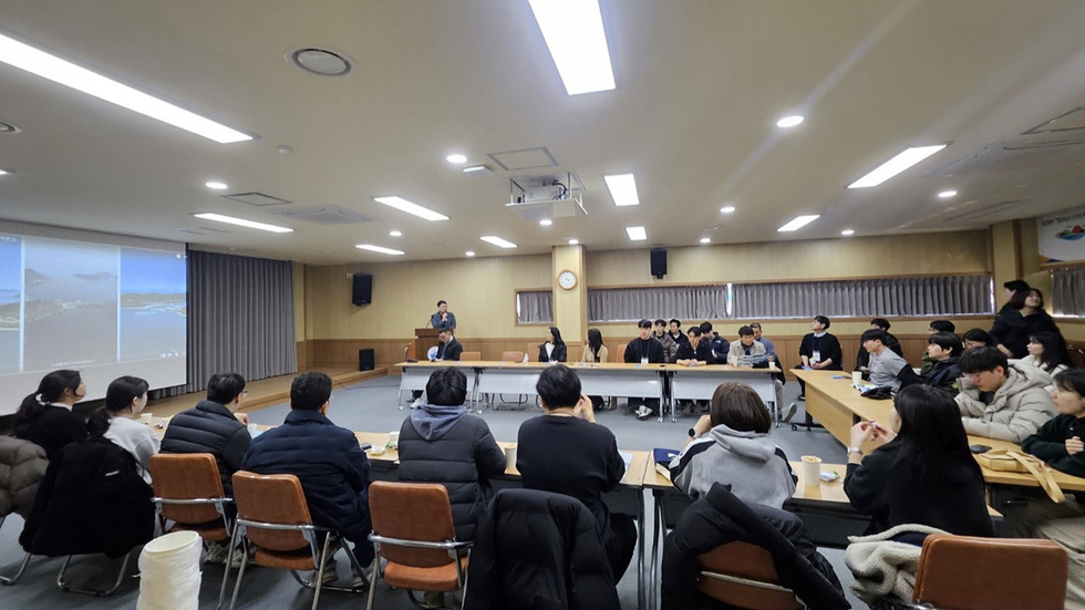 회의실에서 여러 사람이 모여 발표를 듣고 있는 장면이다. 한 남성이 발표를 하고 있으며, 참석자들은 테이블에 앉아 발표를 주의 깊게 듣고 있다.