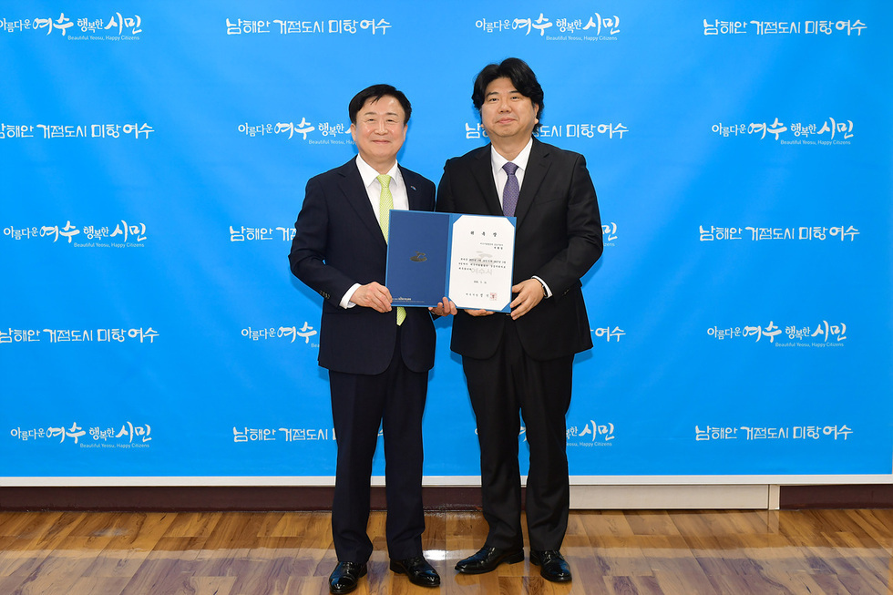 여수시청에서 두 사람이 서류를 들고 기념사진을 찍고 있다. 배경에는 여수시의 슬로건이 적힌 파란색 배너가 있다.