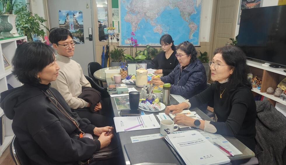 여섯 명의 사람들이 회의 테이블에 앉아 대화하고 있는 모습으로, 테이블 위에는 문서와 음료가 놓여 있다. 배경에는 세계 지도가 걸려 있다.