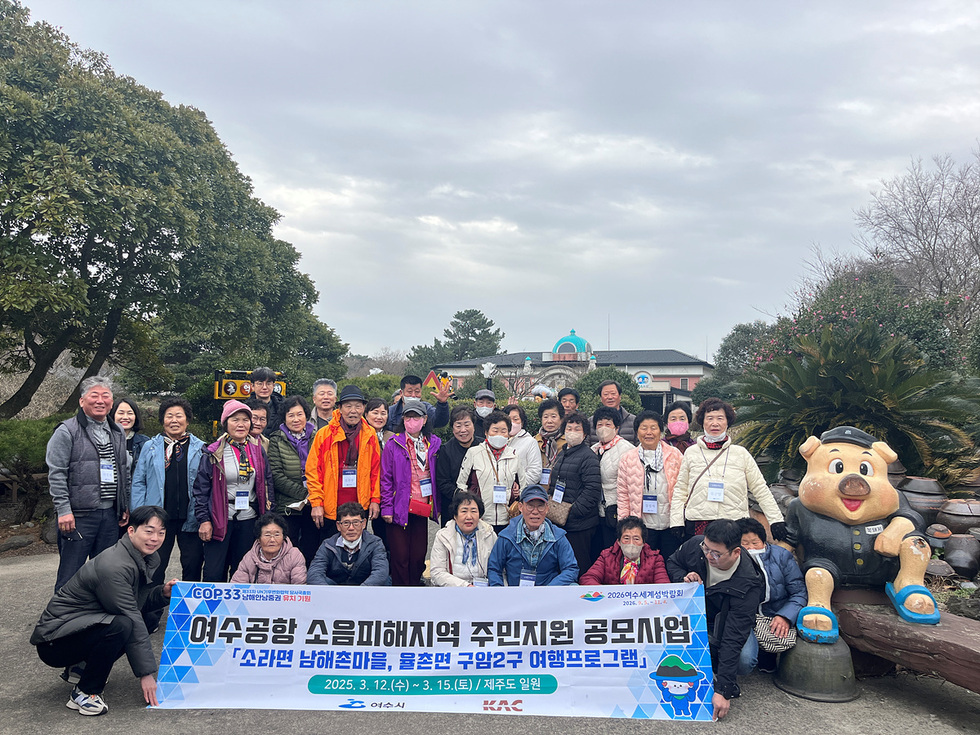 여수공항 소음피해지역 주민들이 여행 프로그램을 위해 모여 사진을 찍고 있는 장면으로, 배경에는 여수시의 건물과 조형물이 보인다.