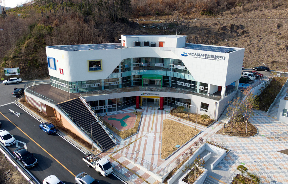 여수시아이돌봄지원센터 건물이 보이는 장면으로, 주변에 차량이 주차되어 있고, 건물 앞에는 정원이 조성되어 있다.