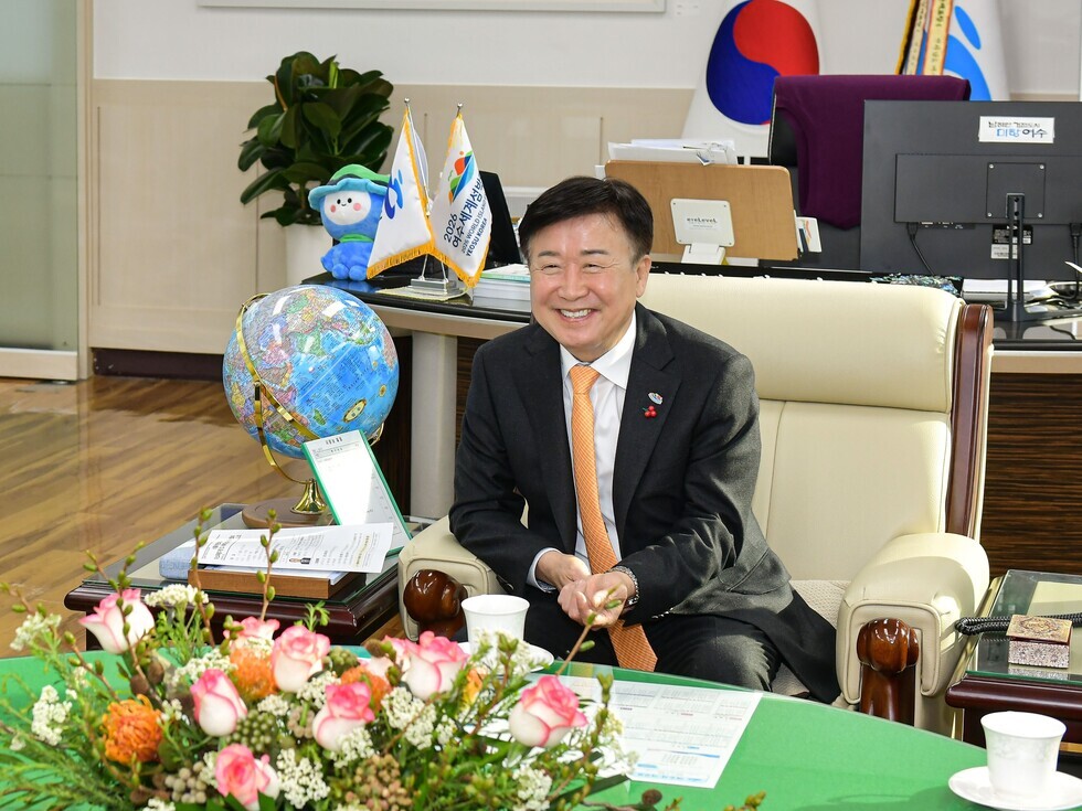 한 남성이 사무실에서 미소를 지으며 앉아 있다. 책상 위에는 지구본과 꽃다발이 놓여 있으며, 배경에는 국기와 여러 가지 사무용품이 보인다.