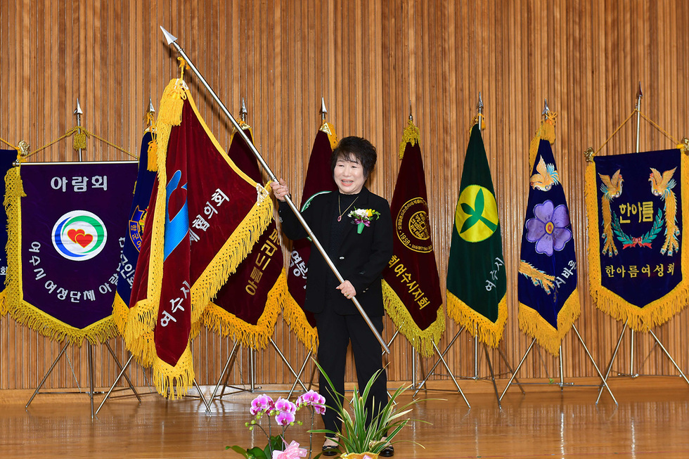 여성단체 아람회의 행사에서 한 여성이 깃발을 들고 서 있다. 배경에는 여러 개의 깃발이 걸려 있다.