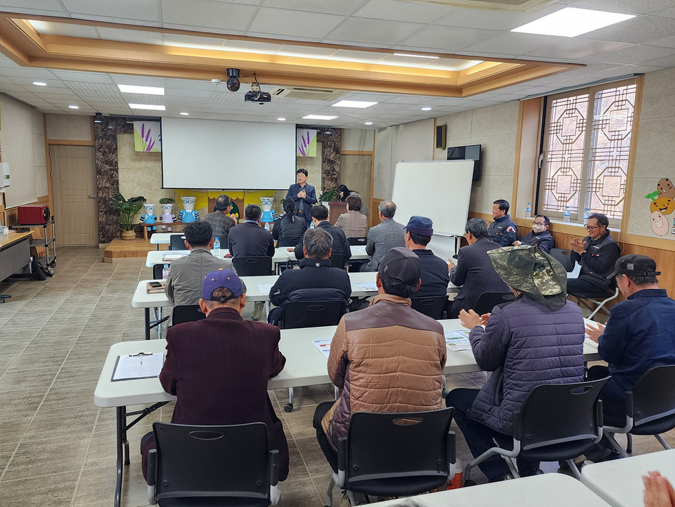 회의실에서 한 남성이 발표를 하고 있으며, 청중은 테이블에 앉아 경청하고 있다. 배경에는 캐릭터 인형과 화이트보드가 있다.