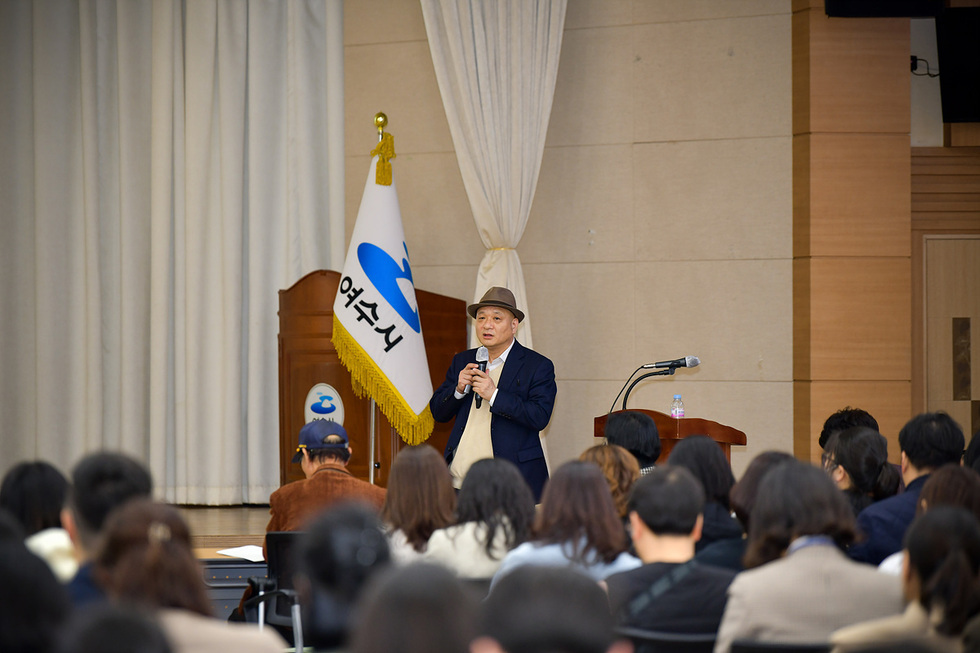 여수시에서 한 남성이 마이크를 들고 청중 앞에서 발표하고 있다. 배경에는 여수시 깃발이 보인다.