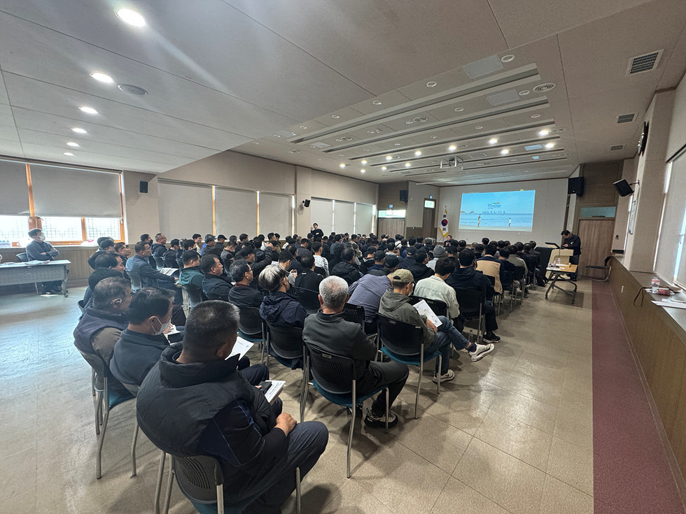 회의실에서 많은 사람들이 발표를 듣고 있는 장면이다. 발표자는 화면 앞에서 설명하고 있으며, 참석자들은 노트와 자료를 참고하고 있다.