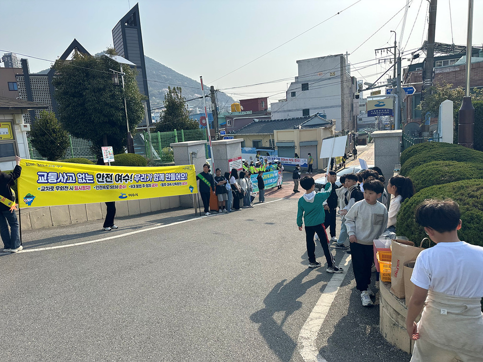 학생들이 교통사고 예방을 위한 캠페인에 참여하고 있으며, 배너를 들고 안전한 여수를 만들자는 메시지를 전하고 있다.