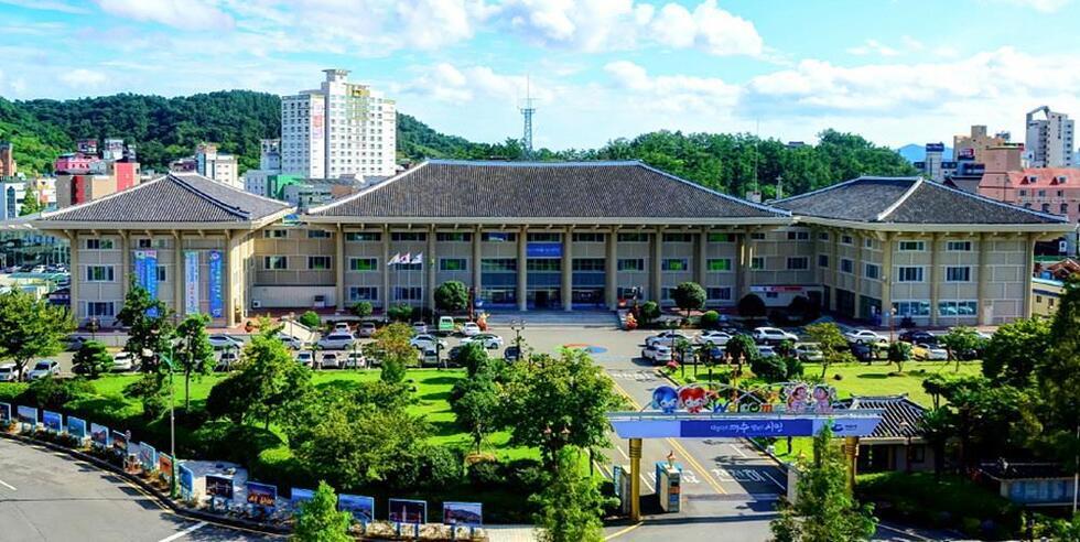 전경에서 보이는 건물은 공공기관으로 보이며, 주변에 나무와 잔디가 조경되어 있다. 건물 앞에는 차량이 주차되어 있고, 뒤편에는 높은 건물들이 있다.