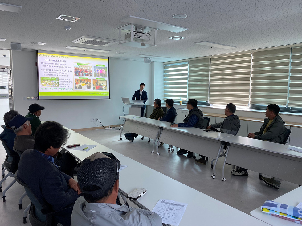 거문도 주민협의체가 회의실에서 강진군 푸소(FU-SO) 운영 사례 강의를 듣고 있는 모습, 앞쪽 스크린에는 푸소 체험 관련 발표 자료가 띄워져 있음