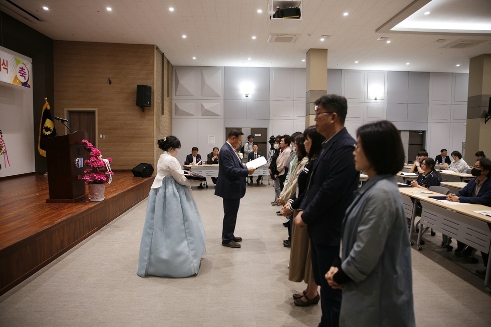 행사장에서 한복을 입은 여성이 남성에게 서류를 전달하고 있으며, 참석자들이 줄을 서서 지켜보고 있는 장면이다.