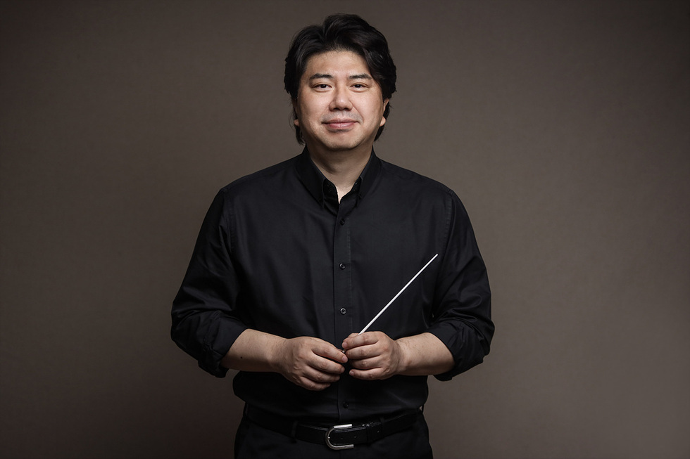 검은색 셔츠를 입은 남성이 지휘봉을 들고 카메라를 바라보며 서 있다.