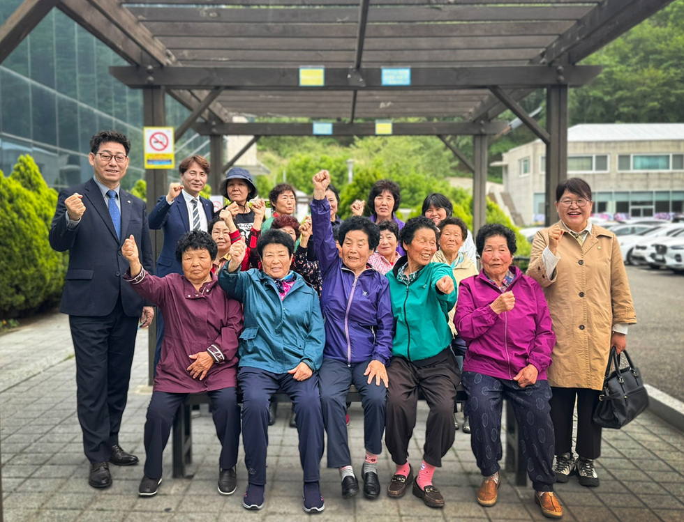 한 남성과 여러 명의 여성들이 야외에서 주먹을 쥐고 포즈를 취하고 있다. 이들은 단체 사진을 찍으며 활기찬 모습을 보이고 있다.