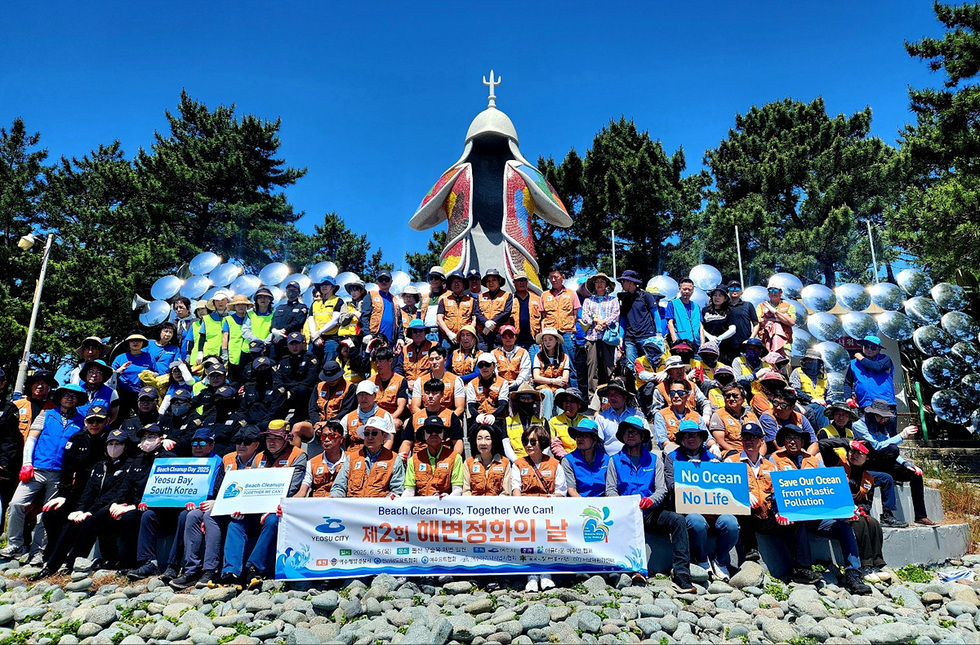 여수 해변 정화 행사에 참여한 사람들로 구성된 단체 사진으로, 다양한 색상의 조끼를 입고 있으며, 배경에는 해양 관련 메시지가 적힌 현수막과 조형물이 있다.