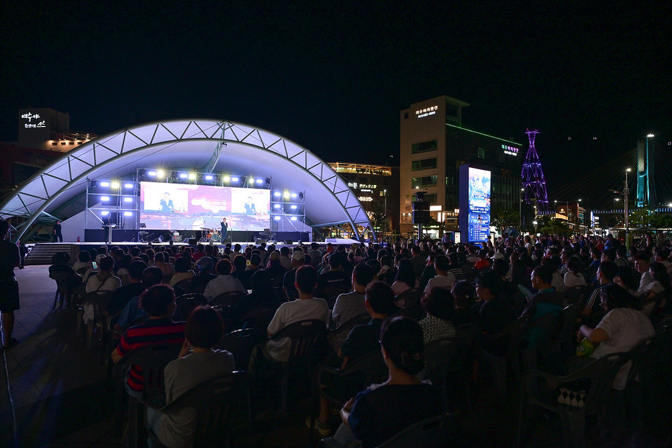 야외 공연장에서 많은 관객들이 무대에서 진행되는 공연을 관람하고 있다. 배경에는 조명이 켜진 건물과 대형 스크린이 보인다.