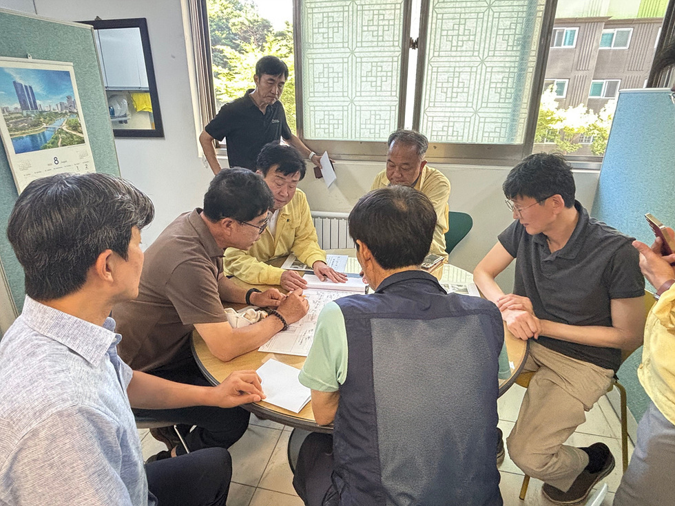 여러 사람이 테이블에 둘러앉아 문서에 대해 논의하고 있는 장면이다. 배경에는 창문과 벽면에 걸린 캘린더가 보인다.