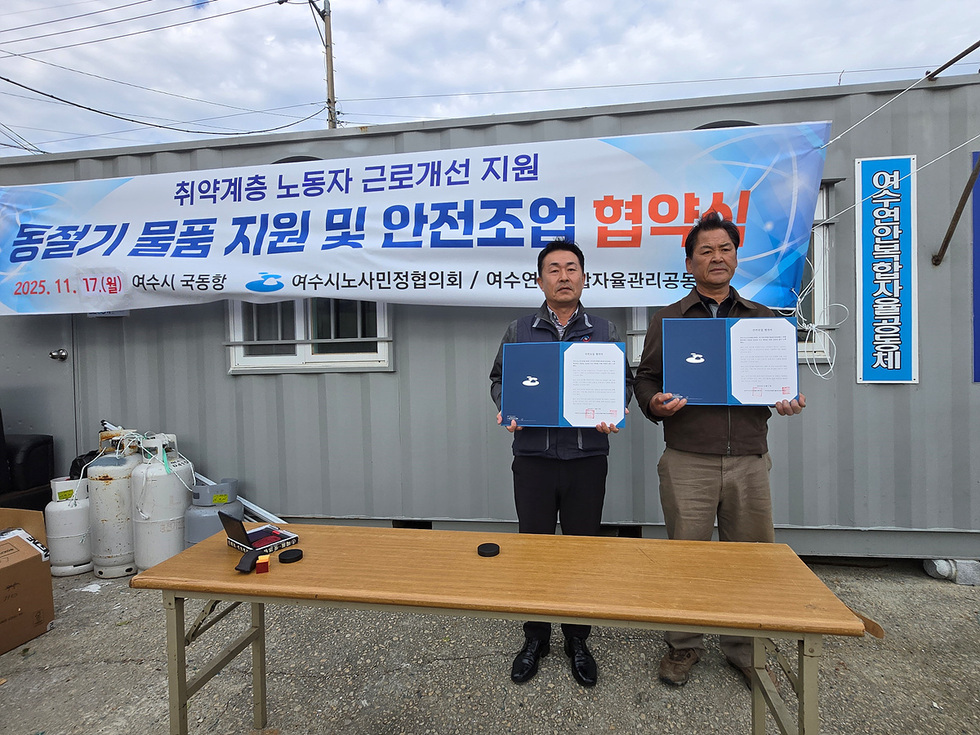 국동항의 임시 컨테이너 사무실 앞에서 두 남성이 협약서 파일을 들고 기념촬영을 하고 있다. 뒤쪽에 ‘취약계층 노동자 근로개선 지원 동절기 물품 지원 및 안전조업 협약식(2025. 11. 17.(월) 여수시 국동항 / 여수시노사민정협의회 / 여수연안복합자율관리공동체)’ 문구가 적힌 현수막이 걸려 있다