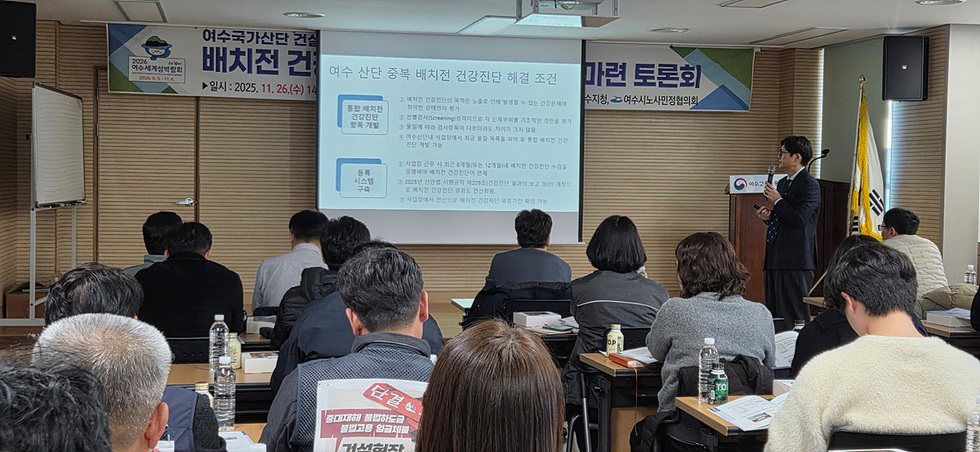 회의실에서 여러 참석자가 앉아 발표를 듣고 있으며, 앞쪽에서 발표자가 배치 전 건강진단 관련 내용을 담은 슬라이드를 설명하고 있다. 화면에는 여수 산단 중복 배치전 건강진단 해결 조건 등이 글자로 표시되어 있다.