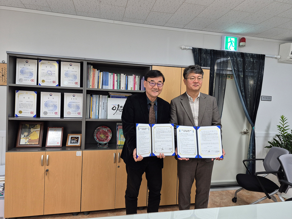 지난 24일 여수시도시재생지원센터와 여수시니어클럽이 2026년도 업무협약을 체결하고 올해에 이어 내년에도 도시재생 공동이용시설 관리와 시니어 사회활동 지원사업을 협업하기로 했다. 사무실에서 두 사람이 협약서를 들고 나란히 서서 기념촬영하고 있다. 뒤쪽 선반에는 각종 인증서와 서적이 진열되어 있다