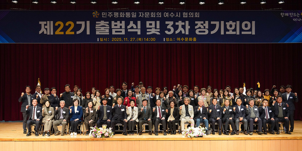 여수문화홀 무대 앞에서 민주평화통일자문회의 여수시협의회 제22기 출범식을 마친 참가자들이 단체로 기념촬영을 하고 있는 모습. 무대 상단에는 ‘제22기 출범식 및 3차 정기회의’라는 현수막이 걸려 있고, 여러 명의 참석자들이 의자에 앉거나 서서 손을 들어 인사하고 있다.