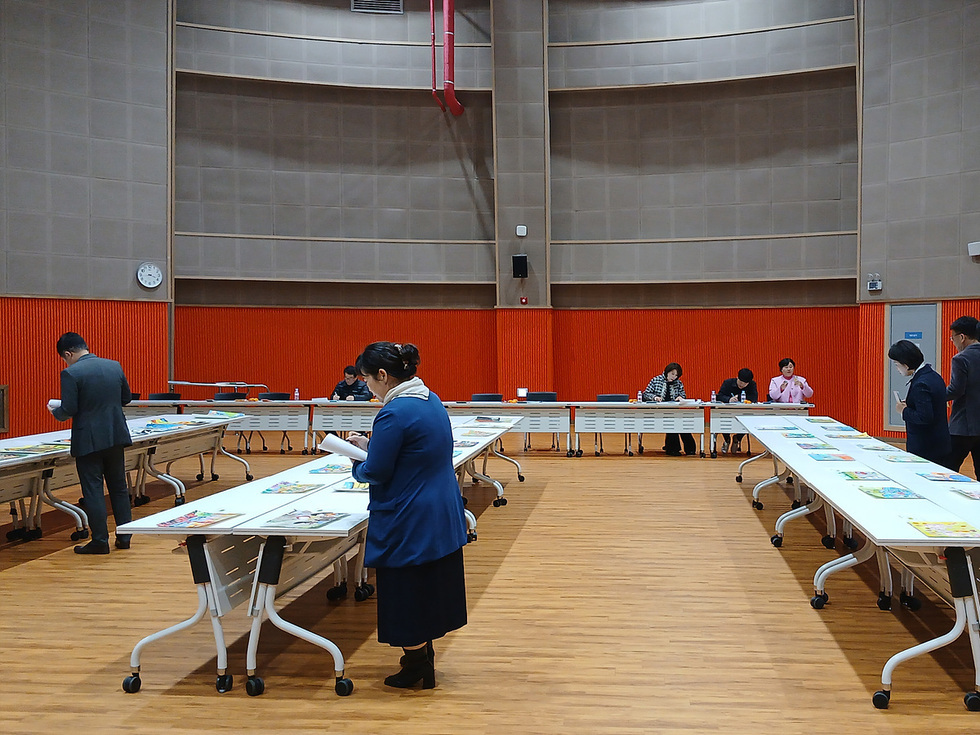 여수시 아동친화도시 그림 공모전 심사가 진행되는 실내 공간에서 심사위원들이 여러 개의 길게 배치된 책상 위에 놓인 공모 작품들을 살펴보고 있다.