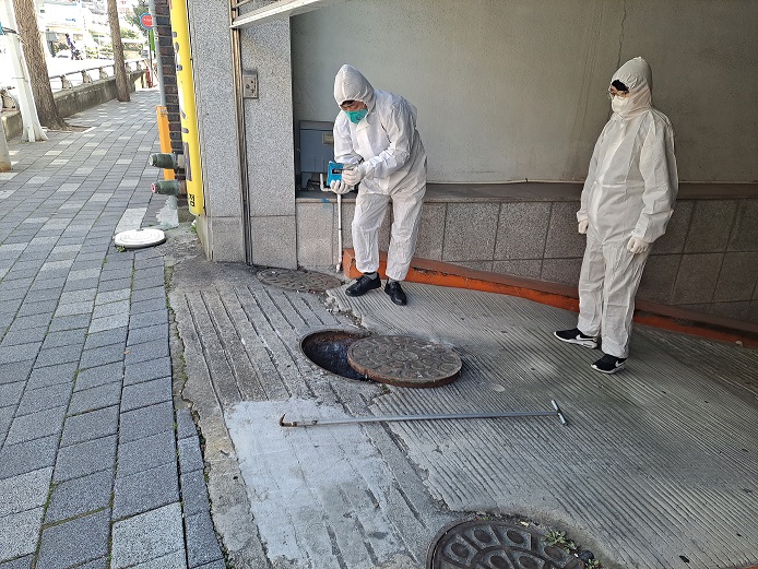 두 명의 작업자가 하수구 근처에서 작업을 하고 있으며, 한 명은 장비를 조작하고 다른 한 명은 지켜보고 있다.