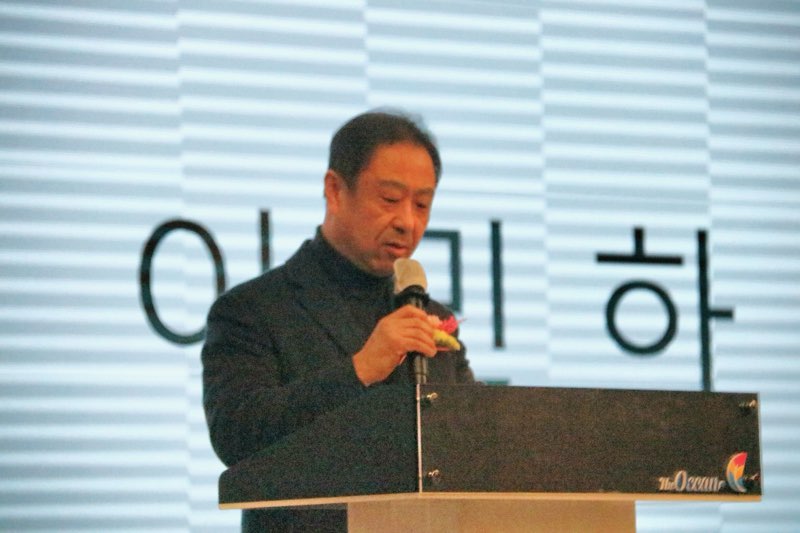 한 남성이 연단에서 마이크를 들고 발언하고 있으며, 배경에는 이 미 하라는 글자가 보인다.