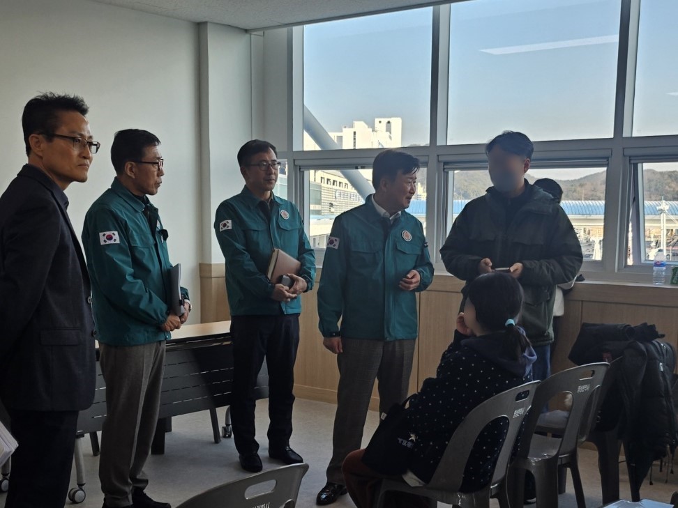 회의실에서 여러 명의 남성이 대화하고 있으며, 한 여성은 뒤쪽에서 이들을 지켜보고 있다. 남성들은 공식적인 복장을 하고 있다.