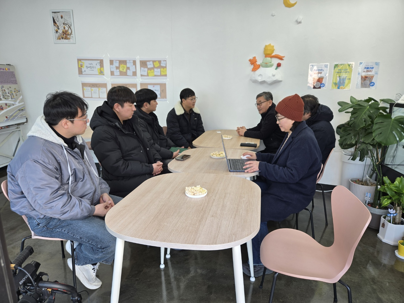 여섯 명의 사람들이 테이블에 앉아 대화하고 있으며, 한 명은 노트북을 사용하고 있다. 배경에는 포스터와 식물이 있다.