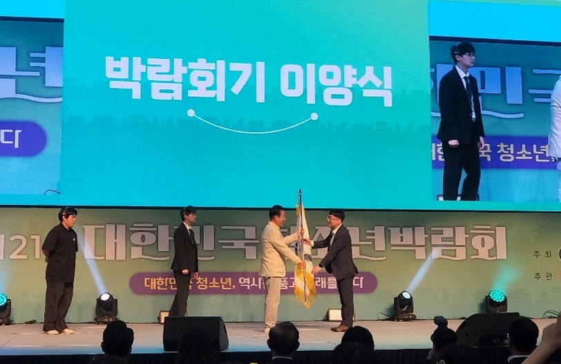 2021 대한민국 청소년 박람회에서 박람회기 이양식이 진행되고 있으며, 두 사람이 기를 교환하고 있다.