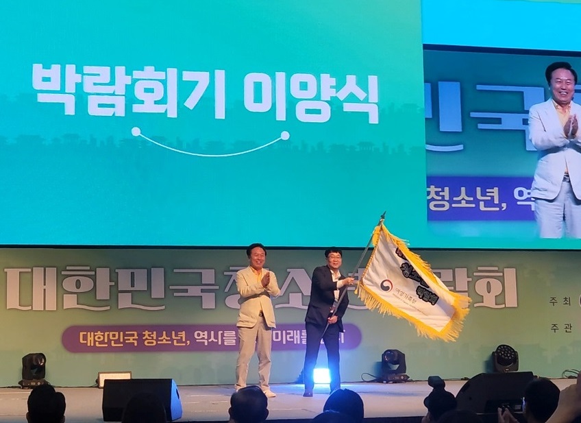 행사에서 두 사람이 무대에 서서 박람회기를 이양하는 장면이다. 배경에는 대한민국 청소년과 역사 관련 내용이 적힌 현수막이 보인다.