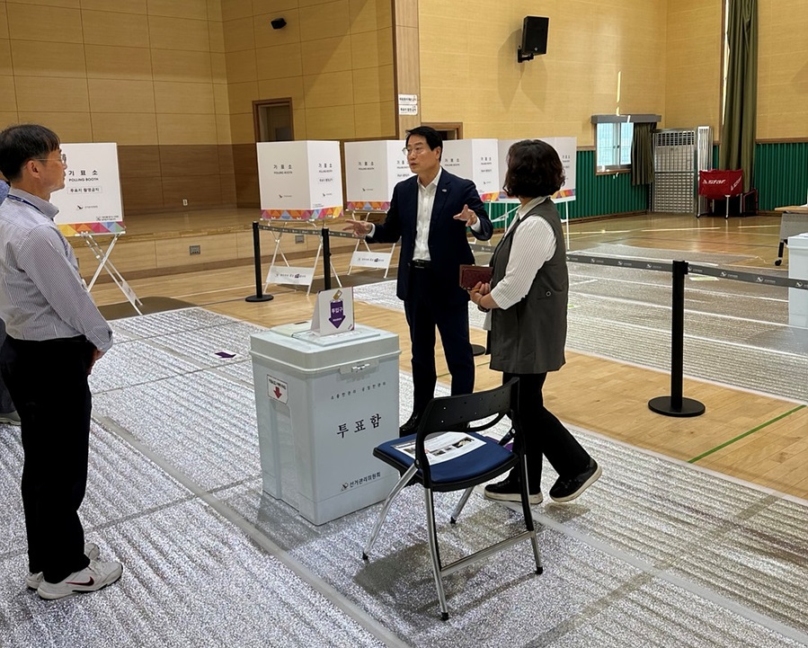 투표소에서 한 남성이 여성에게 설명하고 있으며, 주변에 투표함과 안내판이 보인다.