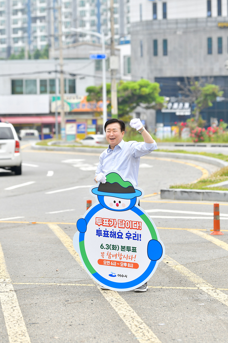 한 남성이 투표를 독려하는 현수막을 들고 도로에서 손을 흔들고 있다. 배경에는 아파트와 상업시설이 보인다.