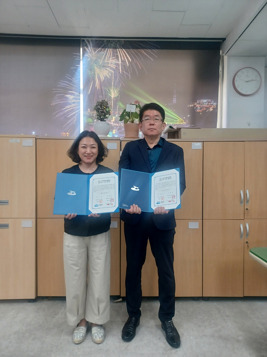 두 사람이 서서 각각 파란색 서류를 들고 있는 모습으로, 배경에는 불꽃놀이와 서울타워가 보인다.