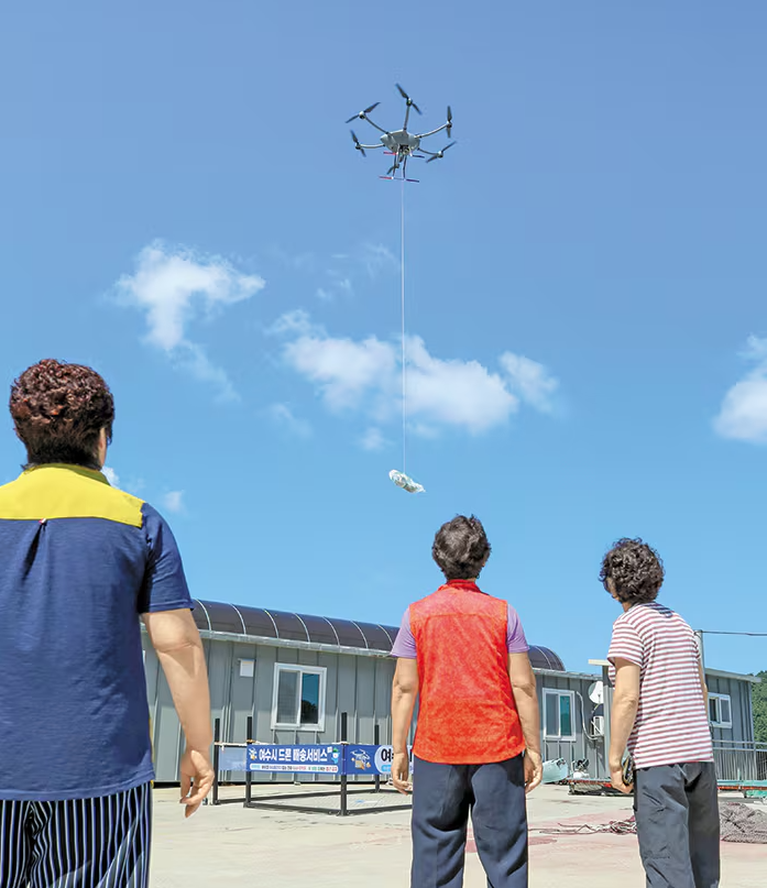 드론이 하늘에서 물품을 매달아 운송하는 모습을 주민들이 지켜보는 장면