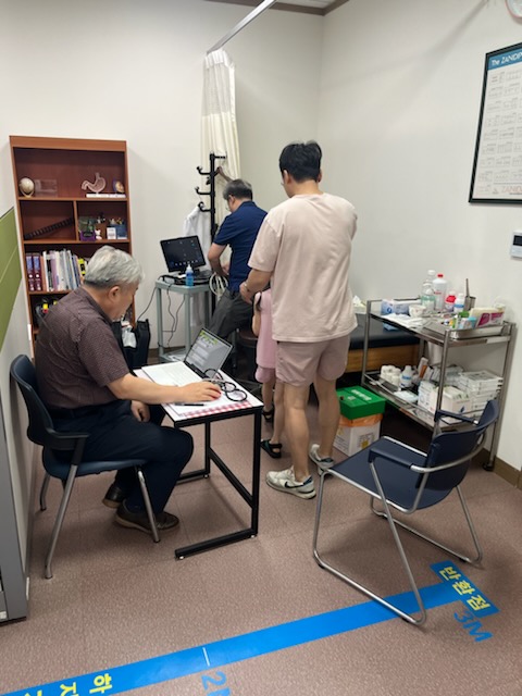 의사와 환자가 진료를 받고 있는 모습으로, 의사는 컴퓨터를 사용하고 있으며 환자는 치료를 받고 있다. 진료실 내부에는 의료 장비와 약품이 보관된 선반이 있다.