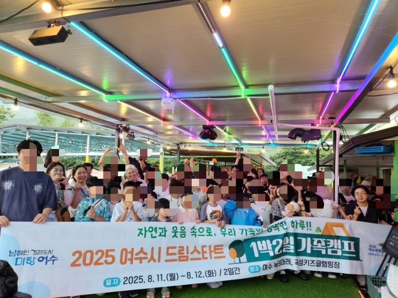 여수시 드림스타트 가족캠프에 참여한 아동과 부모들이 행사 현수막을 들고 단체 기념촬영을 하는 모습, 행사장은 화려한 조명이 설치된 야외 공간