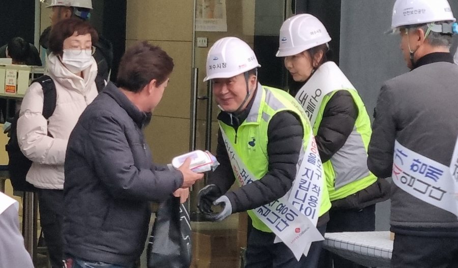 안전모와 형광 조끼를 착용한 정기명 여수시장이 출근하는 근로자에게 응급키트를 건네며 인사하는 모습. 주변에 다른 캠페인 참여자들도 함께 서 있다.